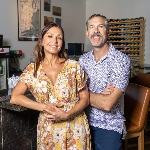 Smiling man and woman standing by a bar with wine racks and sports memorabilia in the background.