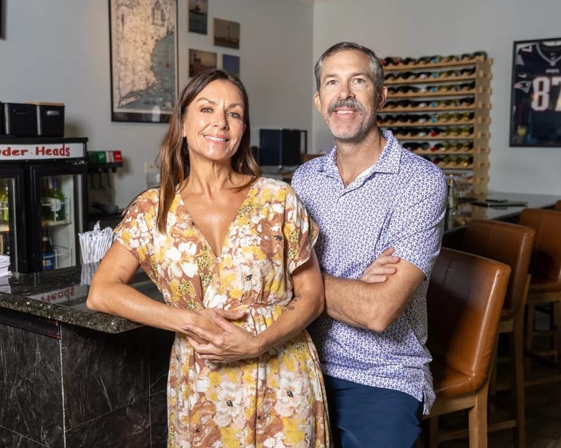 Smiling man and woman standing by a bar with wine racks and sports memorabilia in the background.
