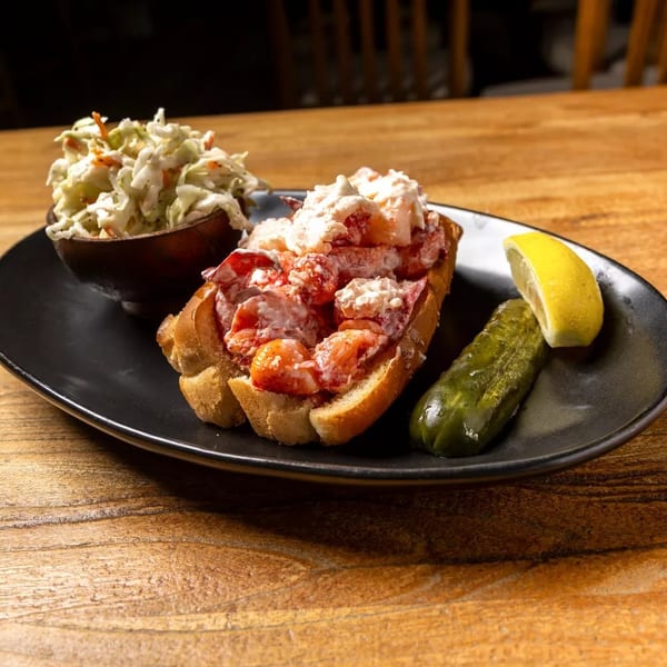 Lobster roll with coleslaw, a pickle spear, and lemon wedge on a black plate.