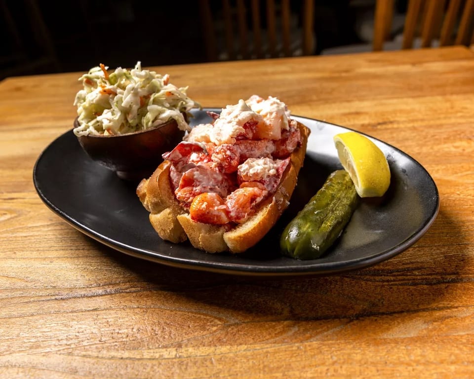 Lobster roll with coleslaw, a pickle spear, and lemon wedge on a black plate.
