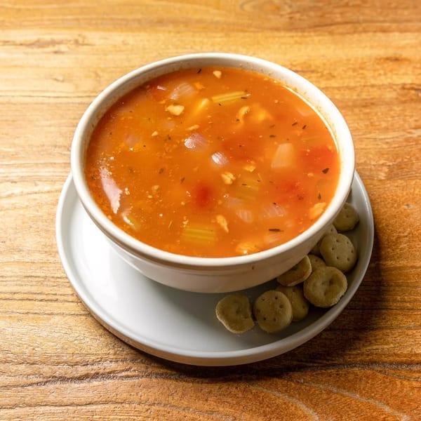 A white bowl of tomato-based soup with vegetables and crackers on a wooden table.