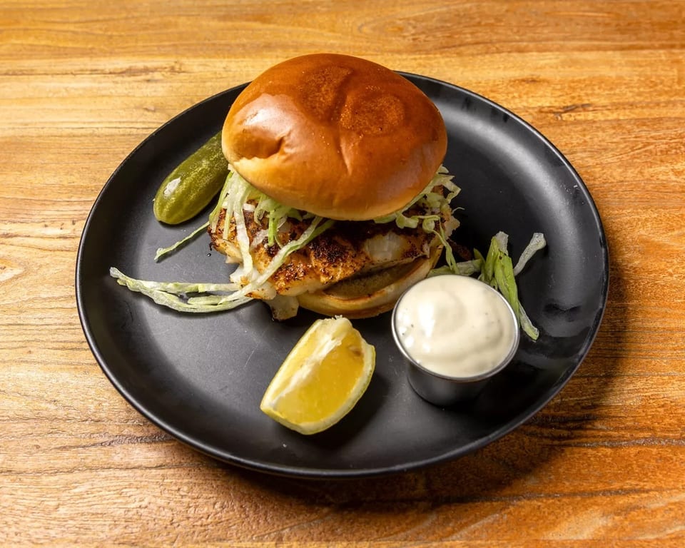Grilled fish sandwich with lettuce, a pickle, lemon wedge, and tartar sauce on a black plate.