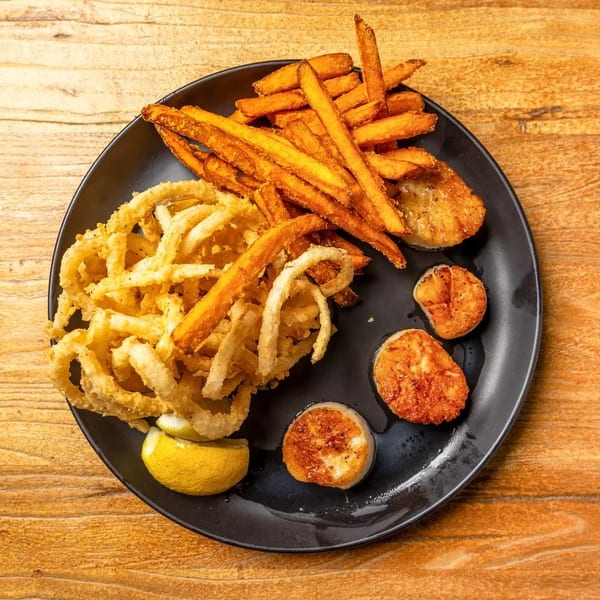 Fried scallops, onion rings, and sweet potato fries with a lemon wedge on a black plate.