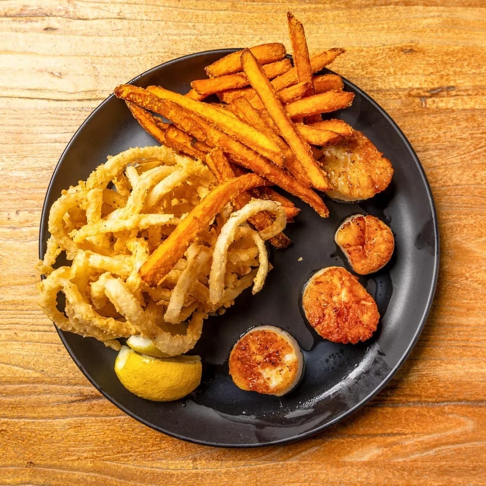 Fried scallops, onion rings, and sweet potato fries with a lemon wedge on a black plate.