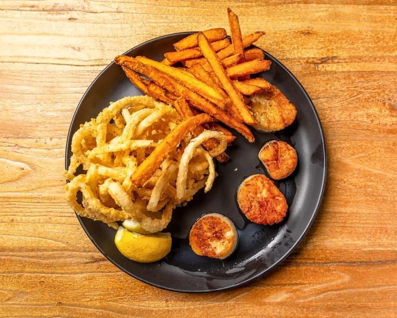 Fried scallops, onion rings, and sweet potato fries with a lemon wedge on a black plate.