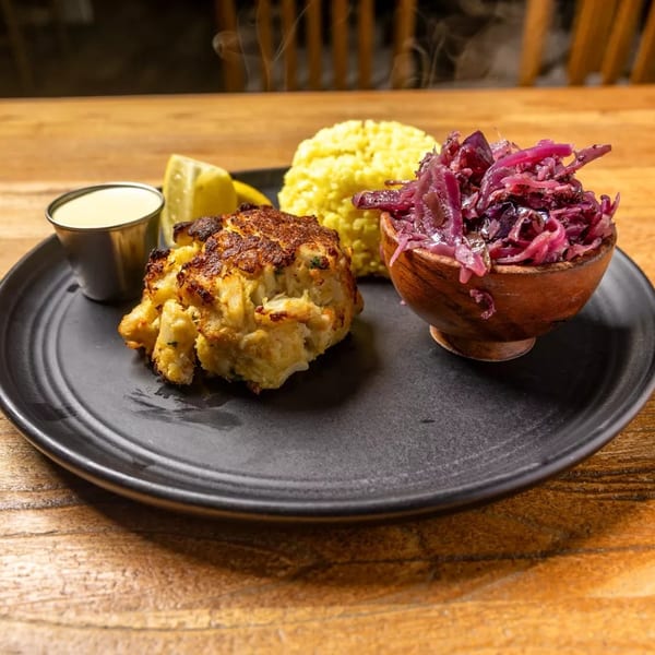 Steaming crab cake, yellow rice, and red cabbage slaw with lemon and sauce on a black plate.