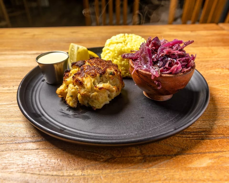 Steaming crab cake, yellow rice, and red cabbage slaw with lemon and sauce on a black plate.